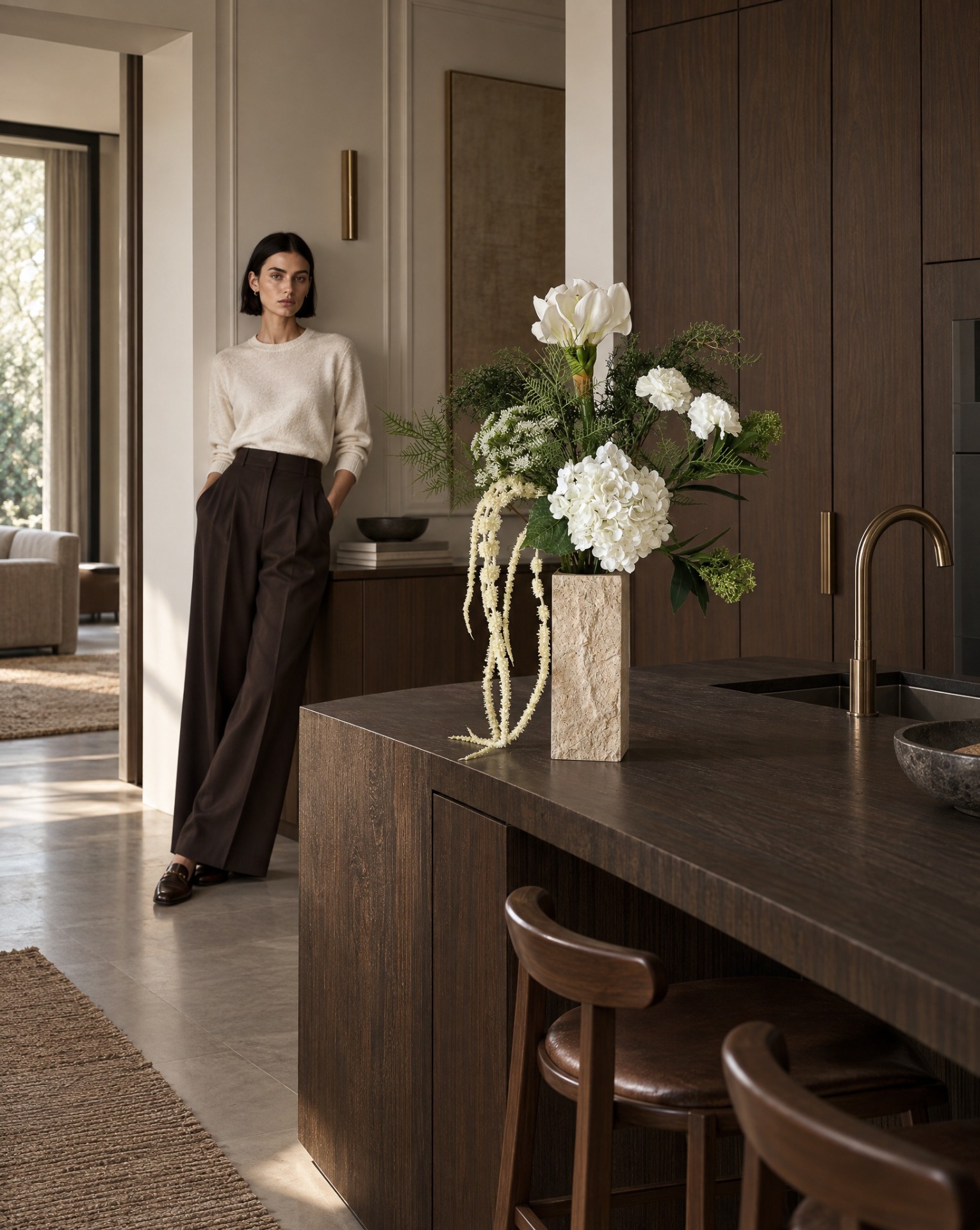 Model staat in moderne keuken, met witte bloemen in vaas op houten aanrecht en gouden kraan.